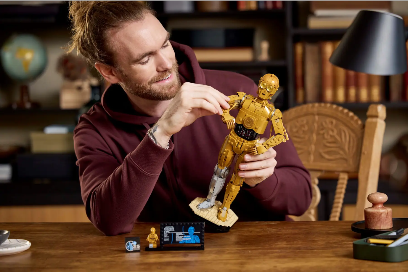 A man in a hoodie assembles a LEGO Star Wars C-3PO model at a desk, with a display stand and mini figures nearby.