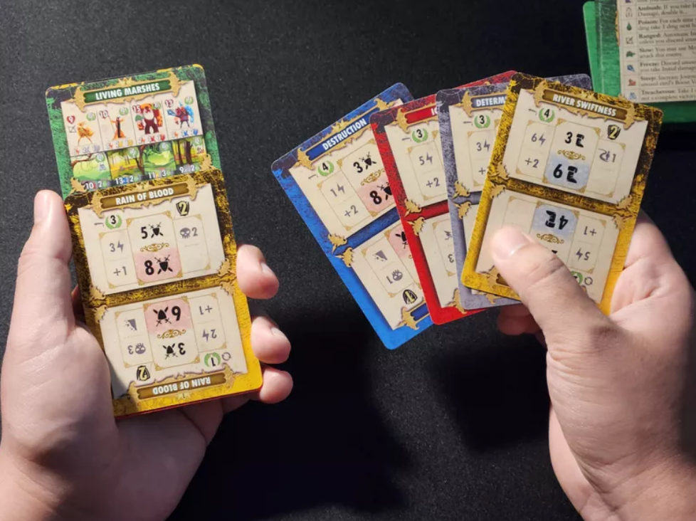 Hands holding game cards from Dragons of Etchinstone, showcasing various stats and abilities for gameplay.