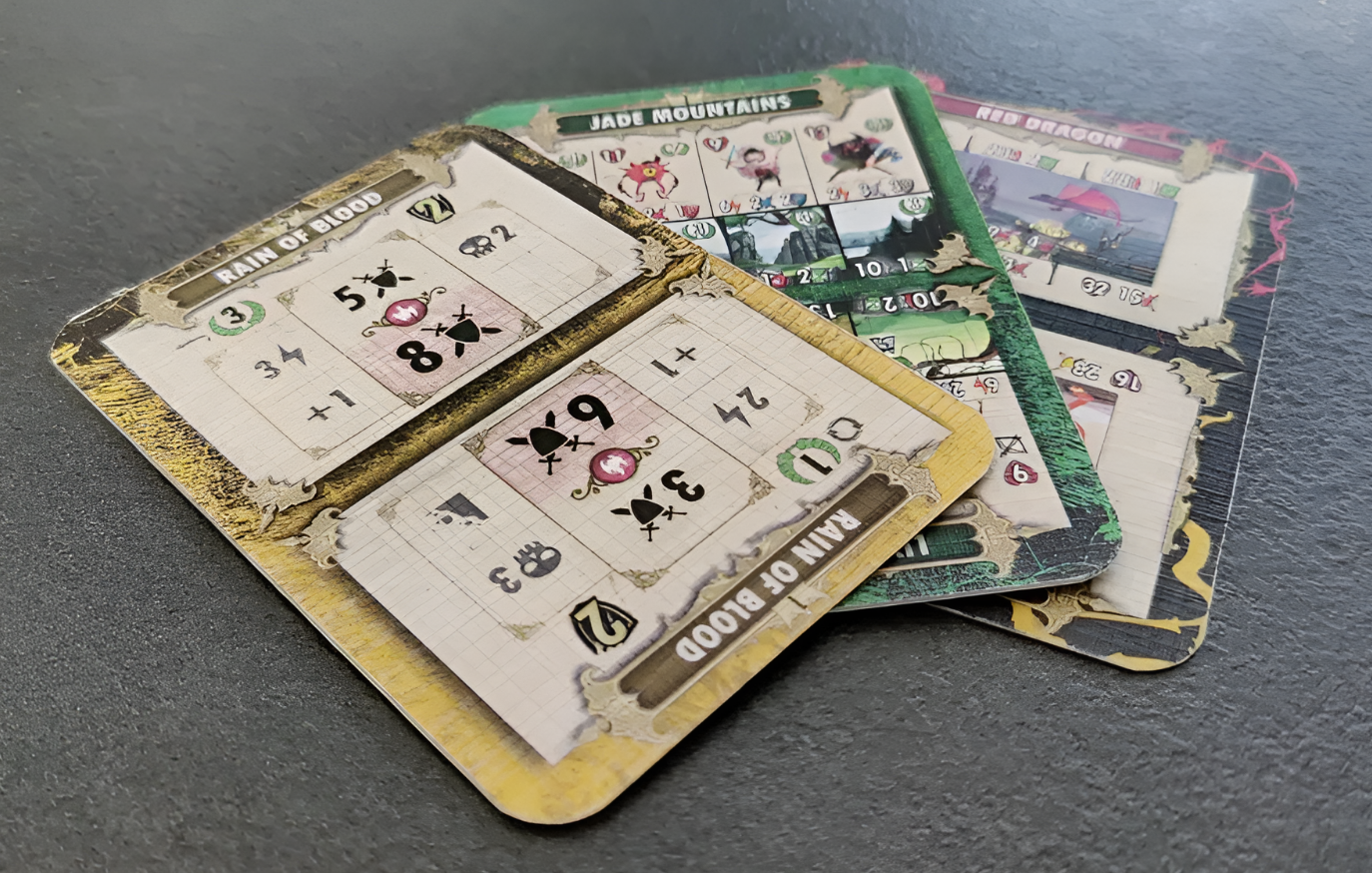 Three game cards from Dragons of Etchinstone featuring stats and abilities for Rain of Blood, Jade Mountains, and Red Dragon.
