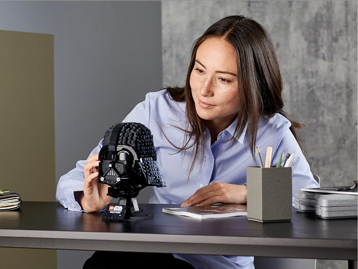 A woman examines the LEGO Star Wars Darth Vader Helmet set on a desk, surrounded by stationery and a notebook.