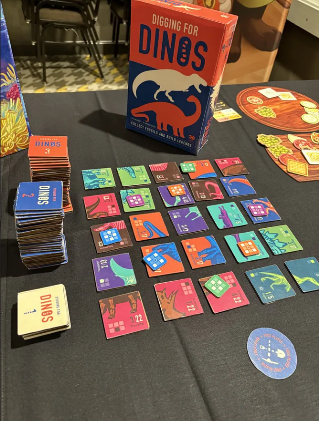 Colorful game setup for "Diggin For Dinos" with cards and tokens displayed on a table, featuring dinosaur-themed artwork.