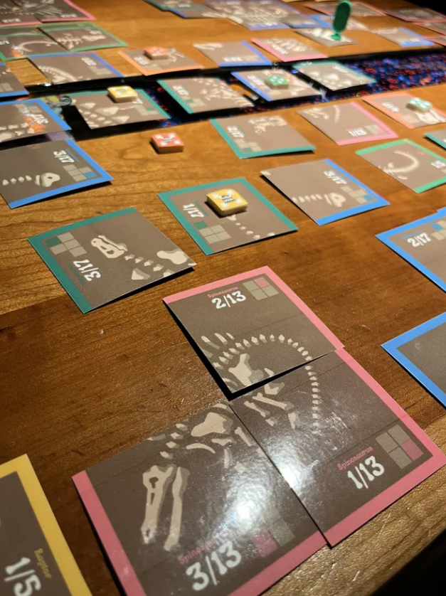 Colorful game cards from Diggin For Dinos laid out on a wooden table, showcasing dinosaur skeletons and game mechanics.