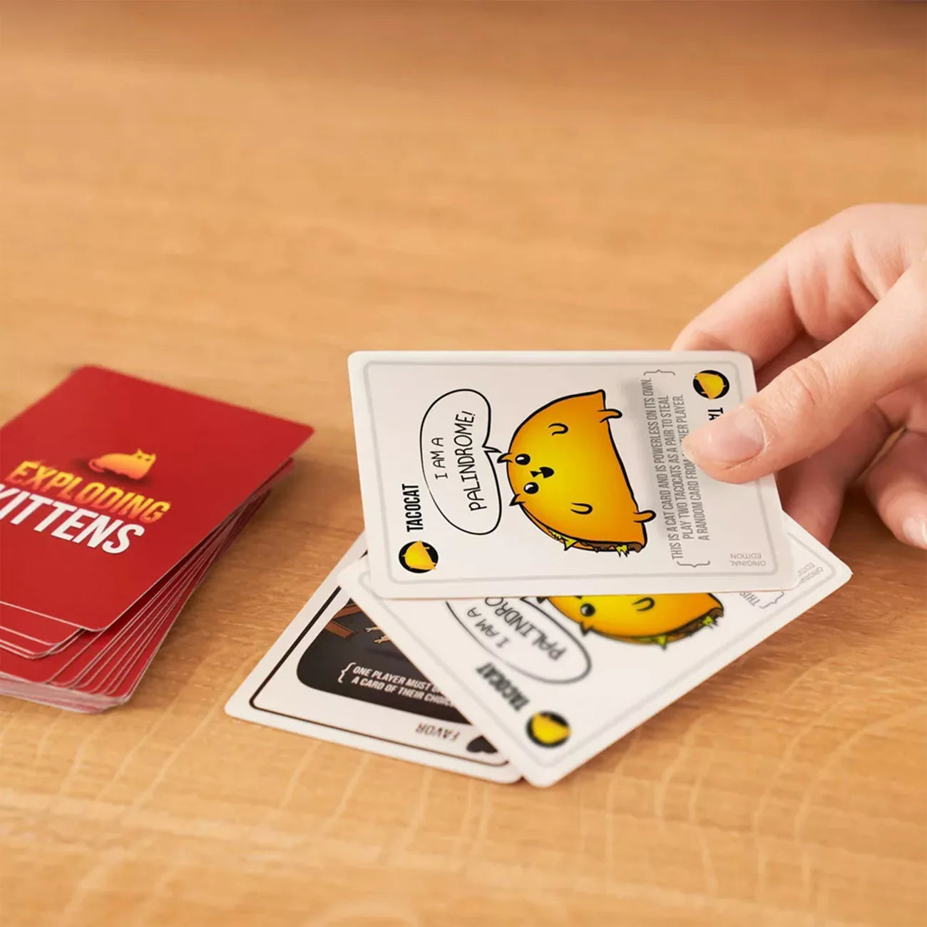 A hand holds an Exploding Kittens card featuring a cartoon cat, with other game cards and the box in the background.