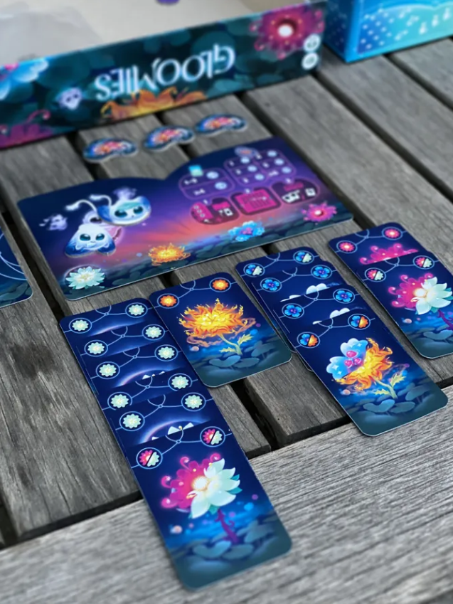 Colorful game components of Gloomies, featuring cards and a game board with whimsical designs on a wooden surface.
