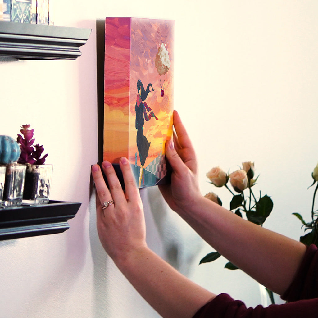 Person hanging a colorful canvas artwork featuring a silhouette against a sunset backdrop on a wall.