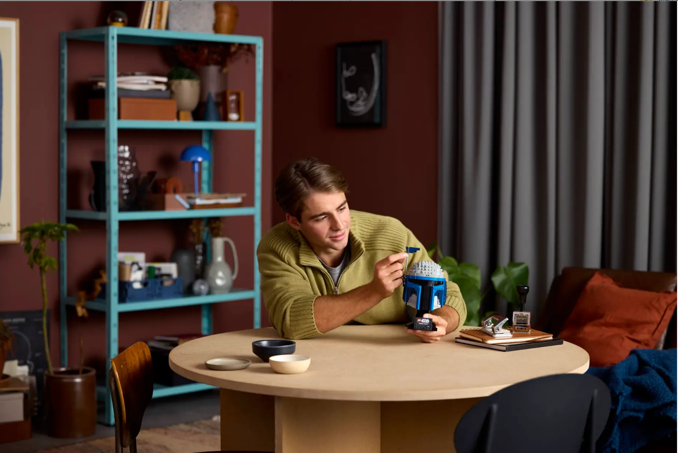 A person assembles the LEGO Star Wars: Jango Fett Helmet (75408) at a table in a cozy room with plants and books.