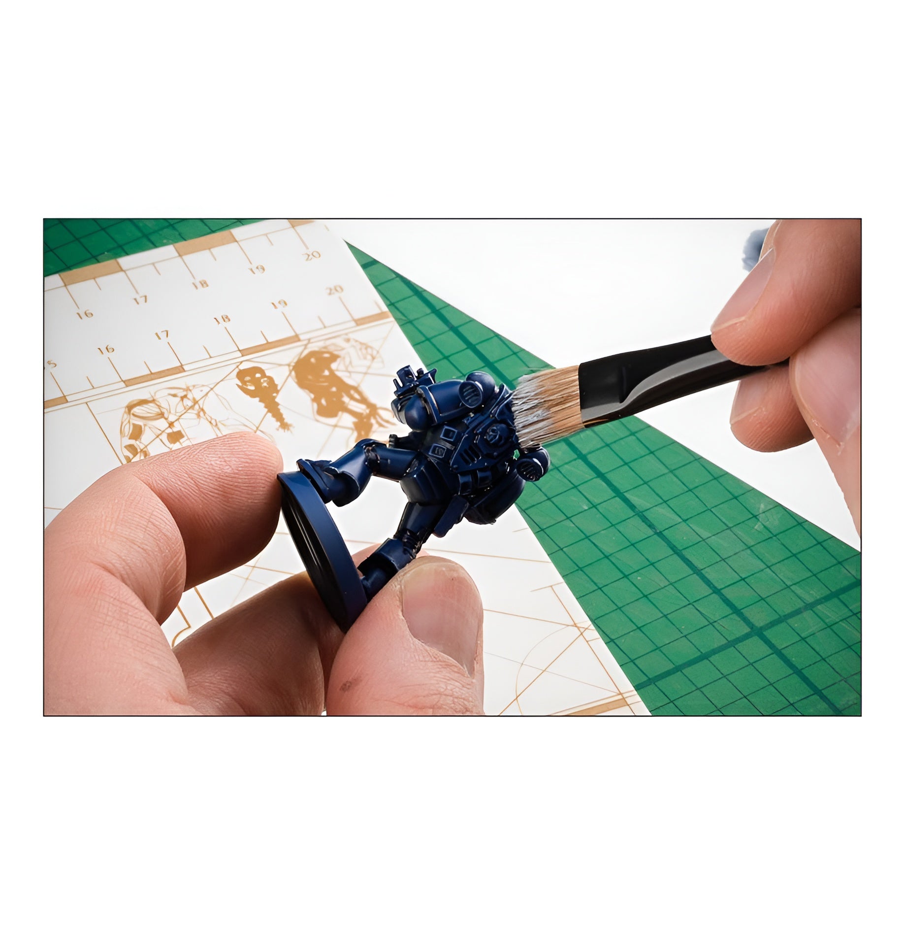 A hand holds a large dry brush applying paint to a blue miniature figure on a green cutting mat.