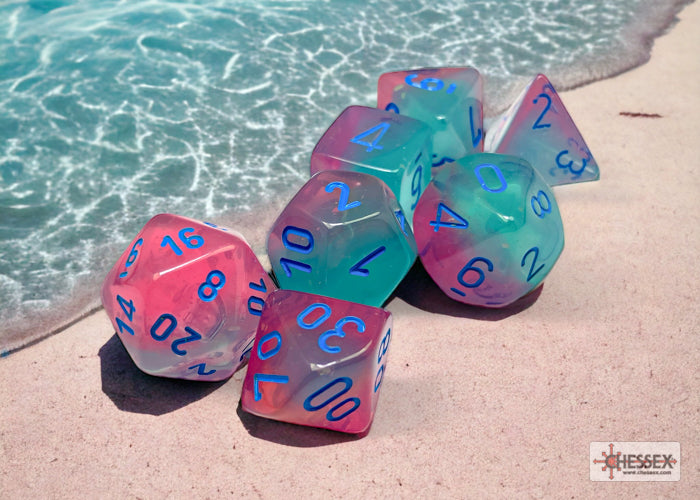 Chessex Gemini Gel Green-Pink/Blue Luminary Polyhedral 7-Dice Set resting on a sandy beach near water, showcasing vibrant colors.