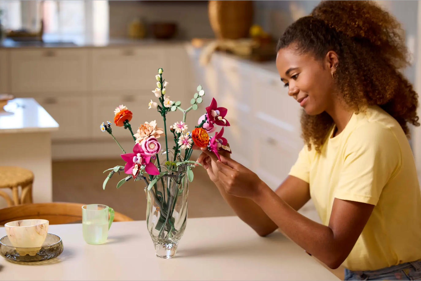 A woman arranges a colorful LEGO flower bouquet in a vase on a kitchen table, showcasing vibrant pink and orange blooms.