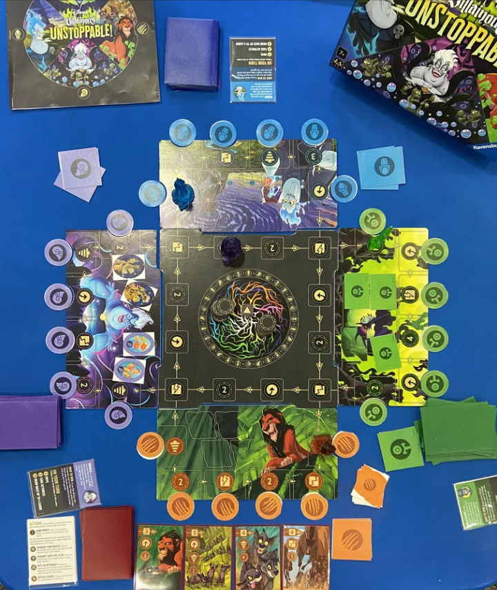 Colorful game board of Disney Villainous: Unstoppable! featuring character cards, tokens, and a box, set on a blue table.