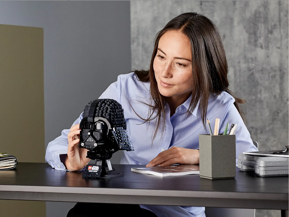 A woman examines the LEGO Star Wars Darth Vader Helmet set on a desk, surrounded by stationery and a notebook.