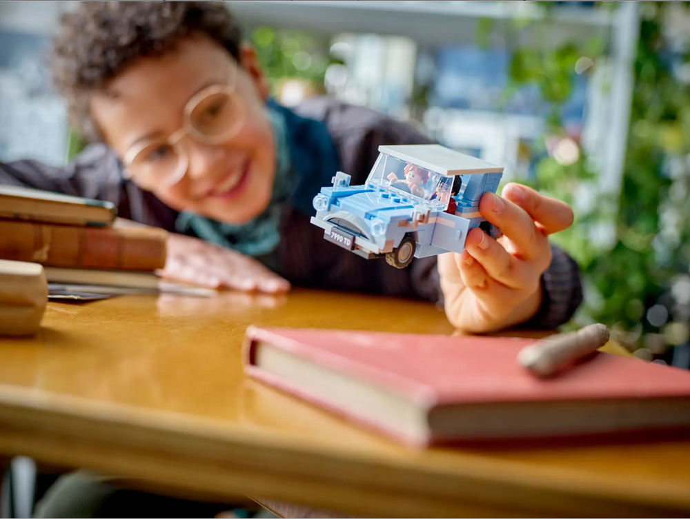 Child joyfully holds a LEGO Harry Potter Flying Ford Anglia model above a table with books, showcasing imaginative play.