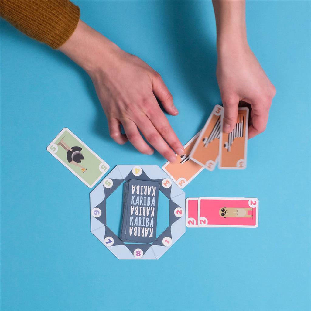 Hands playing the Kariba card game, featuring colorful cards and a central game board on a blue background.