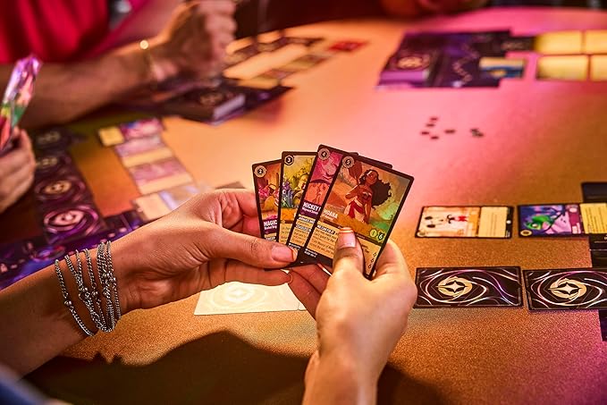 Hands holding four cards from the Disney Lorcana TCG: Into the Inklands, with a colorful game setup in the background.